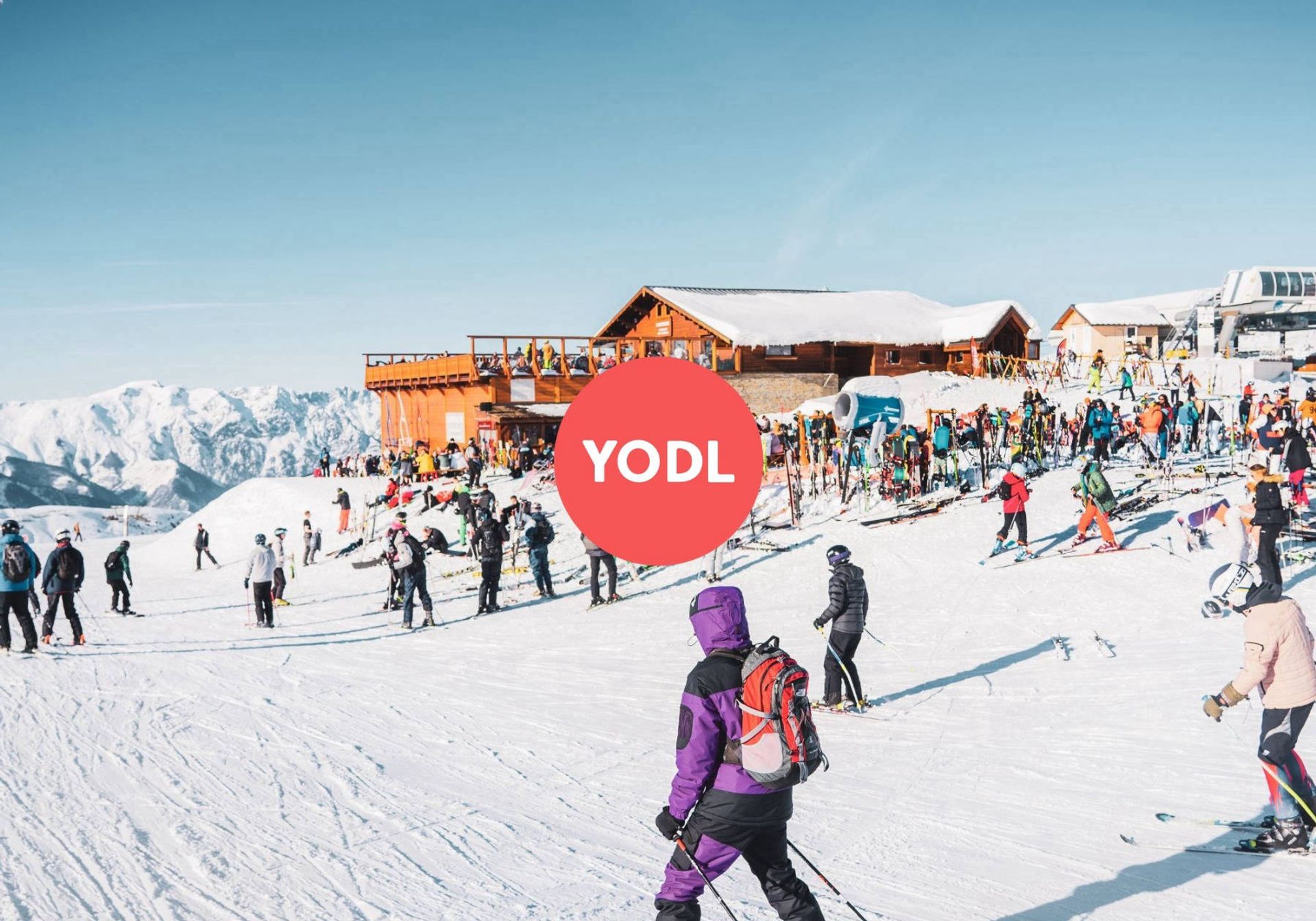 a lot of people in skiing gear standing on a slope with a chalet in the backdrop, a logo of the company 'SkiYodl' is centred in the picture
