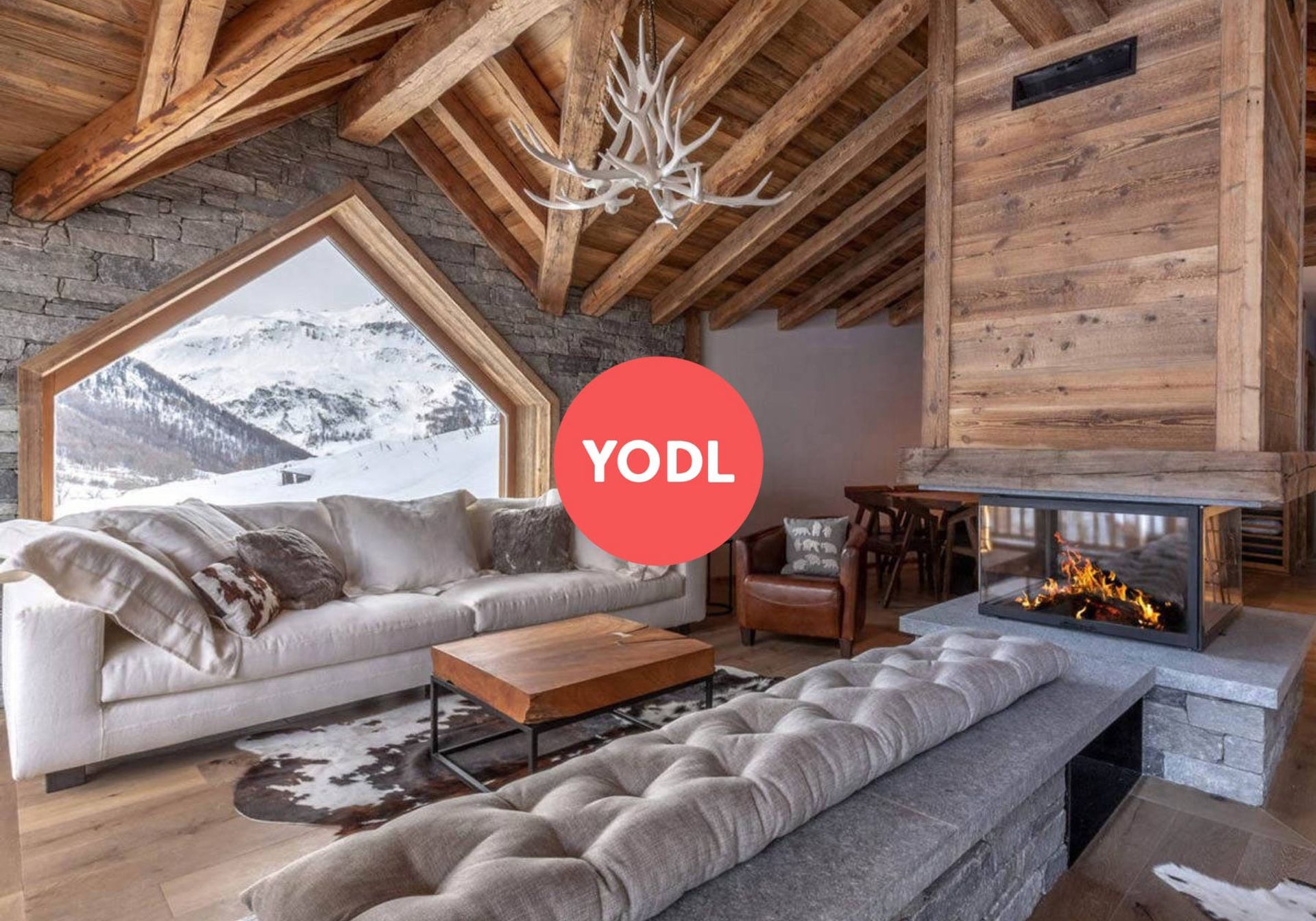 the inside of a chalet living room, with a glass fire place and a burning fire, a white sofa and a large window overlooking the slopes